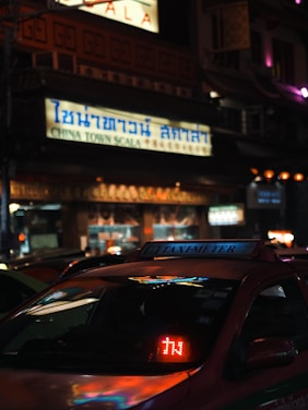 A street scene at night with a taxi in the foreground displaying a light-up 'TAXI-METER' sign. The setting appears to be an urban Chinatown with a large illuminated sign in Thai and English reading 'CHINA TOWN SCALA'. The surrounding area is dimly lit with a mix of colorful lights.