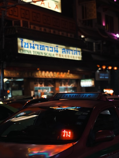 A street scene at night with a taxi in the foreground displaying a light-up 'TAXI-METER' sign. The setting appears to be an urban Chinatown with a large illuminated sign in Thai and English reading 'CHINA TOWN SCALA'. The surrounding area is dimly lit with a mix of colorful lights.