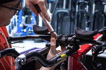 A person wearing glasses and an orange shirt is using a torque wrench to adjust the handlebars on a red bicycle. The background shows various tools and equipment commonly found in a workshop setting.