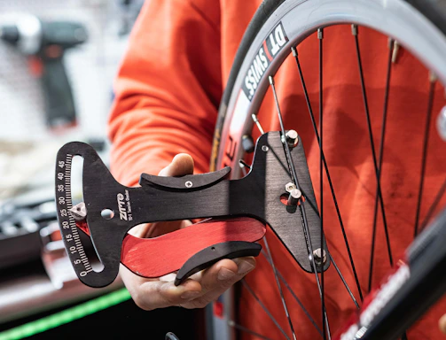 A person is holding a bicycle wheel using a tension meter tool. The tool is placed against the spokes of the wheel to measure tension. The background features a blurred workshop setting.