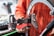 A person is holding a bicycle wheel using a tension meter tool. The tool is placed against the spokes of the wheel to measure tension. The background features a blurred workshop setting.