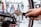 A person is using a wrench to adjust the disc brake on a bicycle wheel. The hand is firmly gripping the handle of the wrench, applying pressure to the bolt. The background is blurred but includes various tools hanging on a perforated wall.
