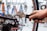 A person is using a wrench to adjust the disc brake on a bicycle wheel. The hand is firmly gripping the handle of the wrench, applying pressure to the bolt. The background is blurred but includes various tools hanging on a perforated wall.