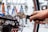 A person is using a wrench to adjust the disc brake on a bicycle wheel. The hand is firmly gripping the handle of the wrench, applying pressure to the bolt. The background is blurred but includes various tools hanging on a perforated wall.