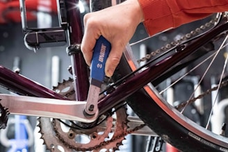 A person in an orange long-sleeve shirt is using a blue-handled tool to work on the crankset of a bicycle. The image focuses on the mechanical components, showing a close-up view of the chain and gears.