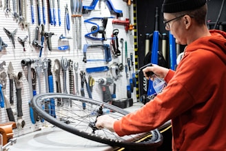 A person wearing a beanie and glasses works on a bicycle wheel, surrounded by an organized display of tools hanging on a pegboard. The person is using a spray bottle, likely for cleaning or lubricating the wheel.