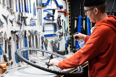 A person wearing a beanie and glasses works on a bicycle wheel, surrounded by an organized display of tools hanging on a pegboard. The person is using a spray bottle, likely for cleaning or lubricating the wheel.