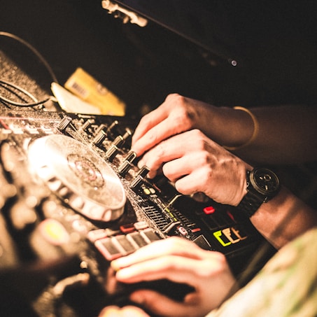 Close-up of hands adjusting controls on a DJ mixer, creating a lively and dynamic scene. The warm lighting accentuates the intensity and focus of the activity.