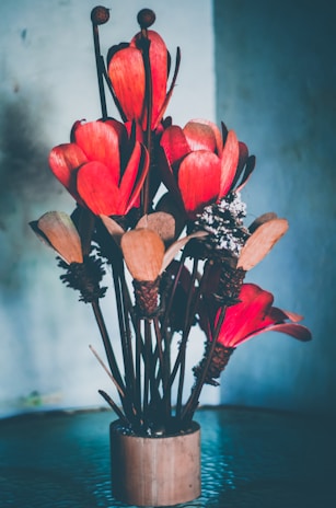 A bouquet of large, red artificial flowers arranged in a brown cylindrical vase. The petals are made of textured material, and the background is softly blurred with gradient shades of blue and gray.