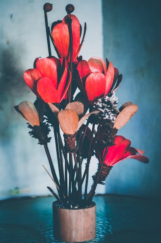 A bouquet of large, red artificial flowers arranged in a brown cylindrical vase. The petals are made of textured material, and the background is softly blurred with gradient shades of blue and gray.