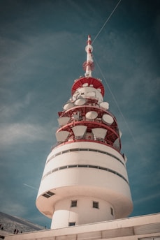 A large, cylindrical communication tower with a red and white color scheme is set against a blue sky. The structure features several satellite dishes and antennas mounted around its upper section. The base of the tower has small windows, and there are cables extending from the top into the sky.