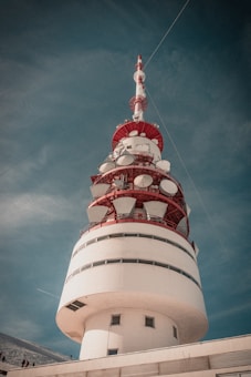 A large, cylindrical communication tower with a red and white color scheme is set against a blue sky. The structure features several satellite dishes and antennas mounted around its upper section. The base of the tower has small windows, and there are cables extending from the top into the sky.