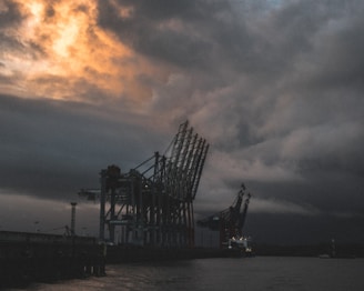 A powerful overhead crane lifting heavy cargo inside a busy industrial port at dusk with dramatic lighting.