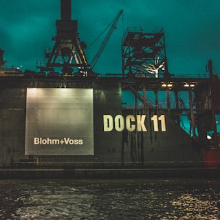 A large industrial dock structure featuring the words 'Blohm+Voss' and 'DOCK 11' prominently displayed. The scene is set against a backdrop of cranes and metal frameworks, with the water in the foreground reflecting the industrial atmosphere.
