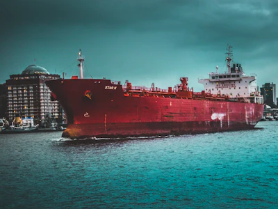 A large red cargo ship labeled 'STAR N' is navigating through a body of water. The vessel is prominently positioned in the foreground, featuring various equipment and structures on its deck. In the background, there are several buildings along the waterfront under an overcast sky, contributing to a dramatic scene.