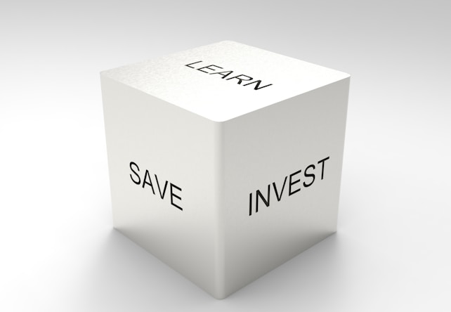 A three-dimensional cube with words on its visible sides, featuring 'LEARN' on the top, 'SAVE' on the left face, and 'INVEST' on the right face. The cube is rendered in a minimalist white and gray color palette, creating a clean and professional look.