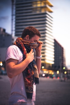 A person wearing a white t-shirt is holding a colorful scarf up to their face. They are standing in front of a tall building with geometric patterns. The background is softly blurred, giving emphasis to the subject. There are lights visible in the distance, suggesting an urban setting at dusk.