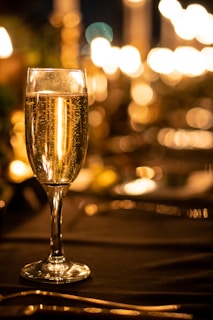 A tall, elegant champagne flute filled with sparkling wine is set on a dimly lit table. The background is softly blurred with warm, glowing bokeh lights, creating a cozy and inviting atmosphere.
