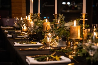 A beautifully set dining table with elegant candles, gold candlesticks, and lush greenery. Rolled white napkins and gold cutlery complement the luxurious atmosphere, while soft lighting enhances a sophisticated ambiance.