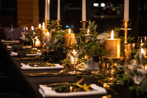 A beautifully set dining table with elegant candles, gold candlesticks, and lush greenery. Rolled white napkins and gold cutlery complement the luxurious atmosphere, while soft lighting enhances a sophisticated ambiance.