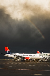 A passenger airplane marked with 'Air Serbia' is stationed on an airport tarmac under a dramatic sky. Dark storm clouds dominate the sky, with a faint rainbow visible in the distance. The airplane is connected to an airbridge as airport staff work nearby.