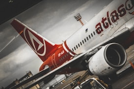 A close-up view of a passenger airplane on the tarmac, with a red and white color scheme and the word 'atlas' partially visible on the fuselage. The airplane appears stationary with one of its engines prominently in the foreground. There is a moody, overcast sky in the background, and part of the airport infrastructure is visible.