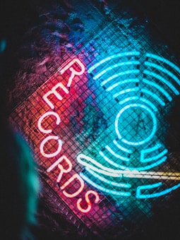 Vibrant neon lights form the word 'RECORDS' alongside a circular, radiating pattern on a textured, dark background. The glowing text is in bright red, contrasting with the blue and teal neon elements that resemble sound waves or records.