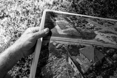 A grayscale image where a hand is holding a photograph. The photograph shows an aerial view of a residential area with a house, a nearby road, several vehicles, and surrounding vegetation. There is a mix of rural and urban elements including what seems to be some clutter or debris near the house.