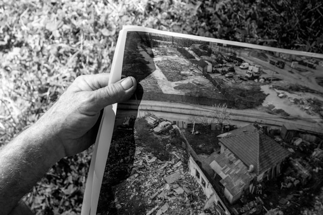 A grayscale image where a hand is holding a photograph. The photograph shows an aerial view of a residential area with a house, a nearby road, several vehicles, and surrounding vegetation. There is a mix of rural and urban elements including what seems to be some clutter or debris near the house.