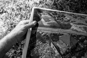 A grayscale image where a hand is holding a photograph. The photograph shows an aerial view of a residential area with a house, a nearby road, several vehicles, and surrounding vegetation. There is a mix of rural and urban elements including what seems to be some clutter or debris near the house.