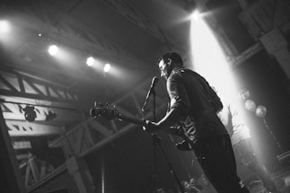 A black and white photo of a musician playing guitar on stage, captured from the side. The stage is lit by several spotlights, creating dramatic shadows. The atmosphere suggests a live music event or concert with an audience in the foreground.