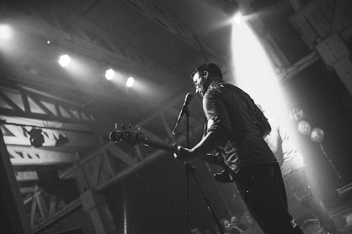 A black and white photo of a musician playing guitar on stage, captured from the side. The stage is lit by several spotlights, creating dramatic shadows. The atmosphere suggests a live music event or concert with an audience in the foreground.