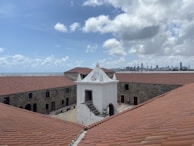 A historic fort with stone walls and a central white structure stands under a partly cloudy sky. Red-tiled roofs cover the surrounding buildings. In the background, a city skyline with high-rise buildings is visible across the water.