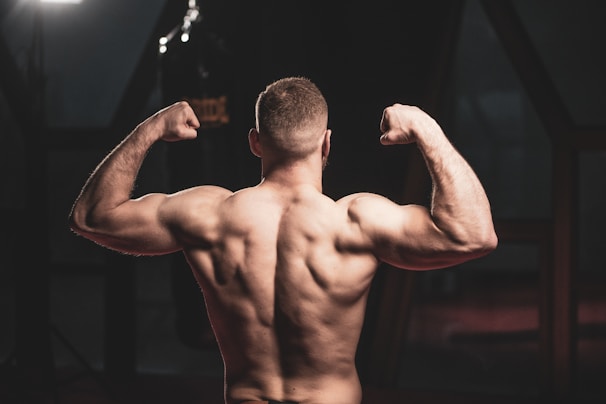 A muscular person is posing with their back facing the camera, flexing their arms to showcase their physique. The setting appears to be a gym or fitness center, with dim lighting creating dramatic shadows and highlighting the muscular definition.