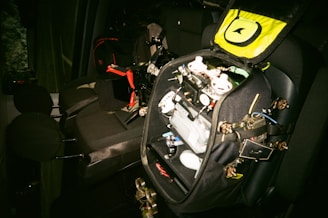 A car interior with a black bag opened on the back seat. The bag contains various electronic components, wires, and possibly a drone frame with propellers. There are black and red details on the bag, and the seats are dark grey.