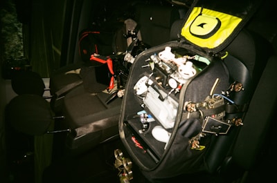 A car interior with a black bag opened on the back seat. The bag contains various electronic components, wires, and possibly a drone frame with propellers. There are black and red details on the bag, and the seats are dark grey.