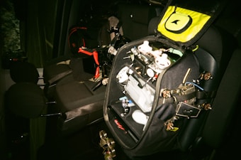 A car interior with a black bag opened on the back seat. The bag contains various electronic components, wires, and possibly a drone frame with propellers. There are black and red details on the bag, and the seats are dark grey.