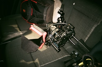 A camera rig with multiple components rests on a car seat. The rig includes a matte box with a red accent and various handles and attachments. A black bag with red details is partially visible next to the rig.