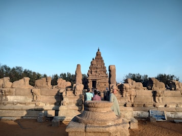 An ancient stone temple is surrounded by sculptures and carvings, with a clear blue sky overhead. Several people are gathered around the temple, some wearing traditional attire. The architecture features intricate designs and historical significance.
