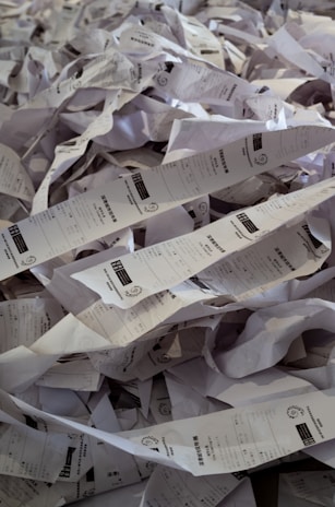 A chaotic pile of crumpled and twisted paper receipts, forming a dense heap. Each receipt has printed text and barcodes visible on some parts. The papers are randomly intertwined, creating a cluttered and messy appearance.