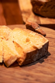 A block of hard cheese with a crumbly texture rests on a wooden board, surrounded by several thin slices. The surface of the cheese is slightly uneven, and its golden hue is contrasted by the dark wooden background.