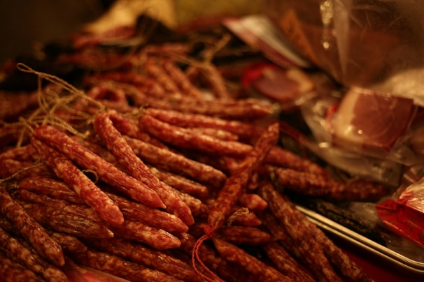 A collection of cured sausages is arranged on a surface, with strings attached for hanging. The background includes packaged meats and has a warm, dim lighting that emphasizes the textures.