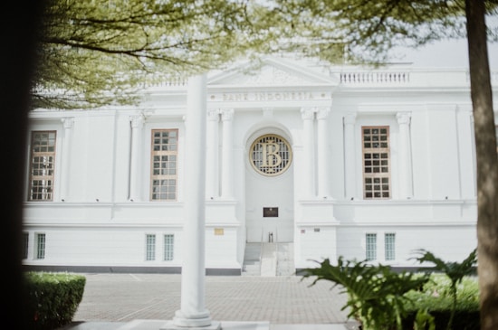 A majestic white building with classical architectural elements such as columns and ornate window frames. The facade features large windows and a central circular emblem. The setting is surrounded by well-maintained greenery and trees.