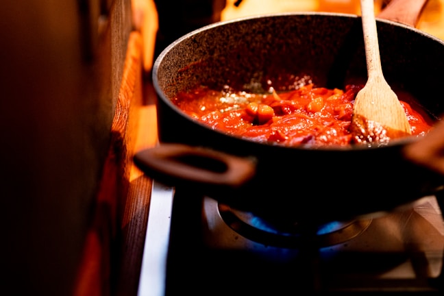 A cooking pan filled with a rich tomato-based sauce being stirred with a wooden spoon. The sauce contains visible chunks of vegetables or meat, simmering gently over a lit gas burner. The kitchen environment appears warm and cozy.