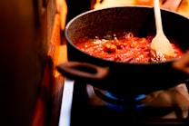 A cooking pan filled with a rich tomato-based sauce being stirred with a wooden spoon. The sauce contains visible chunks of vegetables or meat, simmering gently over a lit gas burner. The kitchen environment appears warm and cozy.