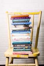 A stack of colorful books is neatly arranged on a worn yellow wooden chair, set against a white brick wall. The books vary in size and color, with titles related to faith, wisdom, and encouragement.