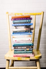 A stack of colorful books is neatly arranged on a worn yellow wooden chair, set against a white brick wall. The books vary in size and color, with titles related to faith, wisdom, and encouragement.