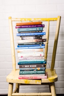 A stack of colorful books is neatly arranged on a worn yellow wooden chair, set against a white brick wall. The books vary in size and color, with titles related to faith, wisdom, and encouragement.