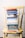 A stack of colorful books is neatly arranged on a worn yellow wooden chair, set against a white brick wall. The books vary in size and color, with titles related to faith, wisdom, and encouragement.
