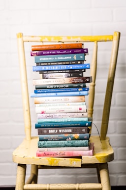 A stack of colorful books is neatly arranged on a worn yellow wooden chair, set against a white brick wall. The books vary in size and color, with titles related to faith, wisdom, and encouragement.